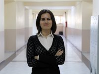 A closeup, waist up, of a female teacher standing in a school hallway. She's looking directly at the camera, her arms crossed, wearing a closed mouth smile. She's wearing a black blouse over a white, collared, button up.