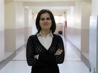 A closeup, waist up, of a female teacher standing in a school hallway. She's looking directly at the camera, her arms crossed, wearing a closed mouth smile. She's wearing a black blouse over a white, collared, button up. 