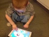A young boy is squatting on a carpet, playing a game on a tablet.