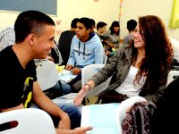 A closeup of two teenage students sitting in a classroom, talking, smiling, and laughing, with other kids sitting and talking in the background.