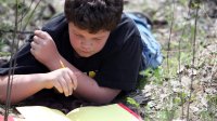 A young boy is lying in the grass with a pencil in his hand, looking down at a red notebook filled with yellow paper
