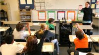 Teacher in front of class pointing to students sitting at desks with raised hands