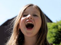 A closeup on a young girl's face, her mouth wide open, singing, with grass and hills in the background.