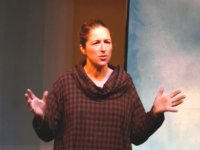 An adult woman with her hair pulled back in a ponytail is wearing a cowl neck maroon and beige checkered hoodie, standing with her elbows at her sides and her hands raised, as if she's addressing an audience. 