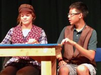 Girl wearing a cap and a boy sitting at a table on stage