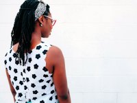 A young woman is standing in front of a white brick wall, her back turned to us at an angle, her eyes closed. 