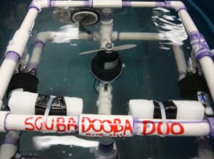 A motorized, square contraption made of plastic pipes is beneath water.