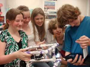 Five students smiling in a classroom, huddling together next to a small computer and a square contraption made of white plastic pipes.