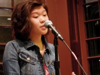 A young teenage girl with shoulder-length hair and a blue jean jacket is standing next to a microphone stand with the microphone up to her mouth. She's standing in the corner of a room with two glass windows behind her.