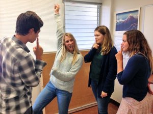 Four students standing together, one striking a pose with arm raised and the others holding fingers to their lips to shhhh