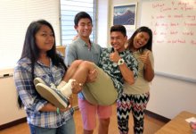 Three teenage students in a classroom, standing side by side, are holding another student and giving a thumbs up.