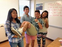 Three teenage students in a classroom, standing side by side, are holding another student and giving a thumbs up.