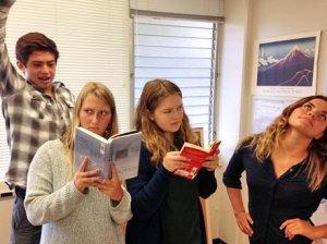 Four students standing together; two are reading while the other two are trying to distract them by striking funny poses