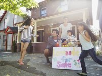Two parents and three children are playing outside on the sidewalk in front of their house, setting up a lemonade stand.