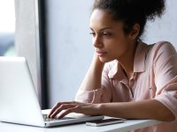 A closeup of a young adult female sitting at a desk, looking at her laptop. She has one hand on the mousepad, and the other is resting against her face with her elbow on the desk.