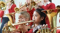 Six young girls in uniform -- a blue skirt, white button up shirt, and a red blouse -- are all playing either the tuba or trumpet.