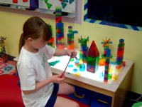 A young girl with long hair in a white t-shirt and blue shorts is sitting at a kid-sized table with colorful, clear, Lego-like building pieces. On the table are variations of different towers.