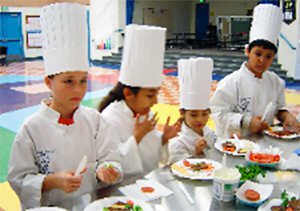 Four kids wearing white chef coats and hats cooking at a long table.