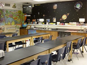 An empty science classroom with three, long tables and a space backdrop against one wall and a dinosaur backdrop against the adjacent wall.