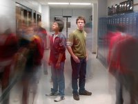 Two students are standing back to back in the middle of the school hallway. Other students are walking past them, blurred, as if to capture all the students walking through the hallway throughout the day.