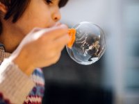 A closeup of a young kid blowing a big bubble. He's to the left of the photo, the top of his head out of frame, and his arm is in front of his face holding an orange plastic device to blow bubbles.