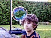 A young boy wearing a striped blue and orange t-shirt is looking at a bubble in front of his forehead that is almost the size of his head. He's outside. Behind him is grass next to a brick floor with a basketball hoop and a tree fence. 
