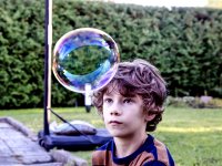 A young boy wearing a striped blue and orange t-shirt is looking at a bubble in front of his forehead that is almost the size of his head. He's outside. Behind him is grass next to a brick floor with a basketball hoop and a tree fence.