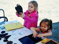 Two young girls are standing on grass next to a table with paper, stamp ink, crayons, and glove-like cloth that they're using as stamps. One girl is pushing her glove into the stamp ink, and the other girl is holding up her glove.