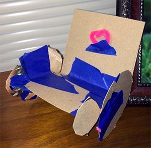 A doll-sized, cardboard wheel chair is on a wooden table. The parts are connected by purple duct tape and pink pipe cleaners.
