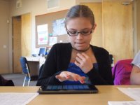 A young girl is sitting at a table in a classroom, working on a tablet. 