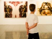 A young boy in a white t-shirt is standing in a museum, with his hands behind his back, looking at artwork. 