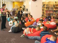A group of about 20 young children are sitting and laying down on bean bags in a library. They're listening to a teacher standing in from of them and talking to them.