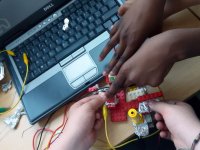 A closeup of two pairs of hands making something out of legos and tin foil over a desk, laptop, and red, yellow, and black wires.