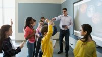 A male teacher and eight young students are standing in a blue-walled classroom near a projector with planets displayed on it.