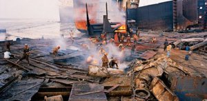 Many men wearing hard hats looking through large piles of metal in a shipyard