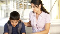 A female adult touching the shoulder of a seated male child who has a sad expression.