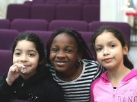 Three young girls are standing side-by-side; the girl in the middle has her arms around the other two. They're all smiling, looking directly at the camera. Behind them are rows of purple theater seats.