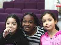 Three young girls are standing side-by-side; the girl in the middle has her arms around the other two. They're all smiling, looking directly at the camera. Behind them are rows of purple theater seats. 