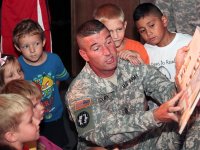 A U.S. army soldier in uniform is kneeling, reading to a group of young kids around him.