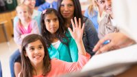 A young girl is raising her hand amidst a group of children and adults sitting together. They're all looking towards the front of the classroom.