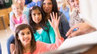 A young girl is raising her hand amidst a group of children and adults sitting together. They're all looking towards the front of the classroom. 