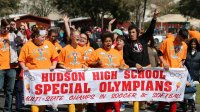 A large group of special needs students are marching in the street, proudly raising their arms, cheering, and holding a large sign that says, "Hudson High School Special Olympians -- Multi-State Champs in Soccer and Softball."