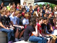 Fifty-three plus teenage students are sitting outside in the sun on raised, cement seating.