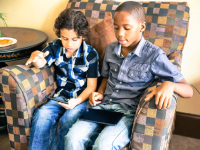 A photo of 2 young boys sitting in a big easy chair, playing games on their tablets.