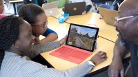 Two girls work in Minecraft in a classroom as a teacher sits with them and talks.