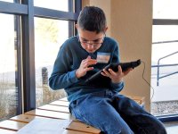 A young boy in a blue sweater and jeans with glasses is sitting on a bench indoors beside a window, holding a tablet and looking at a sheet of paper on the bench.