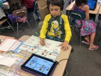 A young boy is sitting at his desk in class in a yellow, long-sleeve Minion shirt. He's wearing headphones connected to a tablet, and is doing math problems on paper that matches what he sees on the tablet.