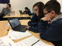 Two students looking at data on a laptop with worksheets scattered in front of them
