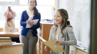 A student confidently speaks to her class as her teacher smiles in the background. 