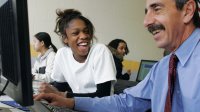 A teacher and student laugh while working at a computer together.